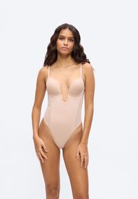 Νεαρή γυναίκα με μακριά κυματιστά μαλλιά, φορώντας κορμάκι σε γυμνή απόχρωση με βαθύ V στο ντεκολτέ, που στέκεται μπροστά σε ένα απλό, φωτεινό φόντο.