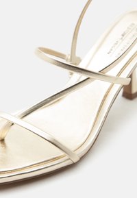 Χρυσό σανδάλι με λουράκια, ανοιχτό δάχτυλο και λεπτά σταυρωτά λουράκια με χαμηλό χοντρό τακούνι σε λευκό φόντο.