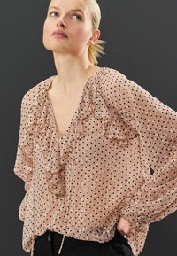 Blouse en tulle rose clair avec des pois noirs, encolure et manches à volants, et ourlet à cordon. Tissu doux et texturé.