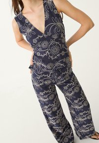 Vrouw in een marineblauwe mouwloze top en broek met witte bloemembellissement, staand met de handen op de heupen tegen een eenduidige achtergrond.