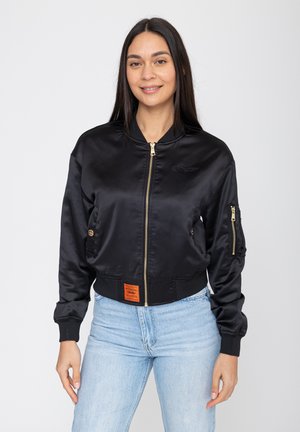 Frau mit langen dunklen Haaren, die eine schwarze Satin-Bomberjacke mit Reißverschluss und hellblaue Jeans trägt, steht vor einem einfarbigen Hintergrund.