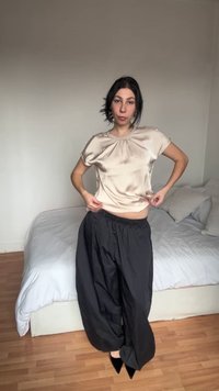 Γυναίκα που προσαρμόζει μια γυαλιστερή μπεζ μπλούζα με κοντά μανίκια, φοράει άνετο μαύρο παντελόνι και μυτερά μαύρα παπούτσια, στέκεται σε υπνοδωμάτιο με κρεβάτι στο παρασκήνιο.