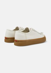 Scarpe in tela color avorio con suola in gomma. Presentano una forma arrotondata, un design minimalista e un'etichetta rossa sul tallone.