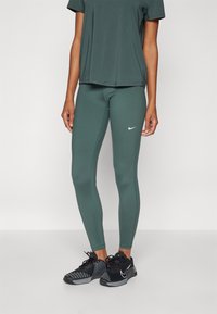 Grønne treningstights med et tætsittende design, med en hvit Nike-logo på siden. Brukt med en matchende løs grågrønn topp og svarte treningssko.