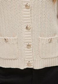 Sweter kardigan w kremowym kolorze z fakturowanym wzorem, z metalowymi guzikami, mankietami ściągaczowymi i dwoma przednimi kieszeniami.