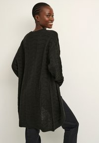 Cárdigan negro de punto extragrande con un patrón texturizado, aberturas laterales y un escote redondeado, llevado sobre pantalones oscuros.