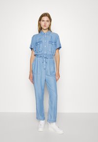 Svetlo modra denim overall z kratkimi rokavi, sprednjim gumbi, elastičnim pasom, stranskimi žepi in zavitimi manšetami, v kombinaciji z belimi supergami.