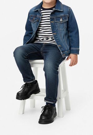 Kind sitzt auf einem weißen Hocker und trägt eine blaue Jeansjacke, ein gestreiftes Hemd, dunkle Jeans und schwarze Schnürschuhe.