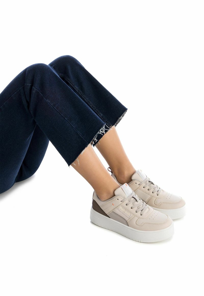 Zapatillas beige con acabado texturizado, con diseño de cordones, acentos en marrón claro y una suela blanca gruesa. Llevadas con vaqueros oscuros recortados.