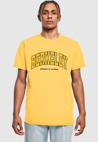 Žluté bavlněné tričko s nápisem "BERKELEY" tučně napsaným černým písmem a nápisem "UNIVERSITY OF CALIFORNIA" pod ním, kulatý výstřih a krátké rukávy.