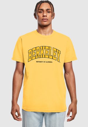 Žlté bavlnené tričko s nápisom "BERKELEY" v tučných čiernych písmenách a "UNIVERSITY OF CALIFORNIA" nižšie, okrúhly výstrih a krátke rukávy.