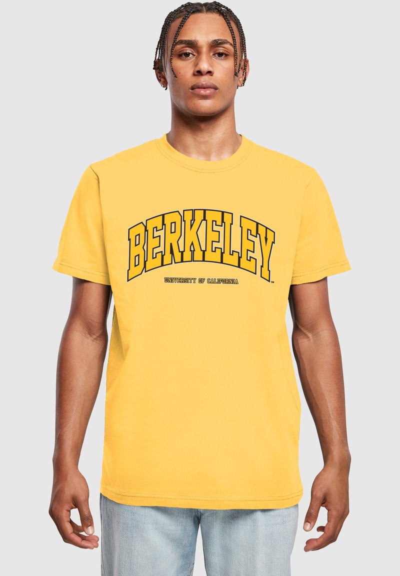 Žluté bavlněné tričko s nápisem "BERKELEY" tučně napsaným černým písmem a nápisem "UNIVERSITY OF CALIFORNIA" pod ním, kulatý výstřih a krátké rukávy.