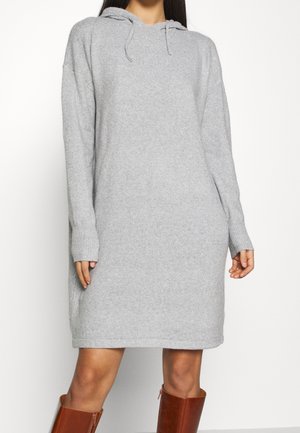 Kobieta ubrana w jasnoszary, długi sweter z kapturem z dzianiny, w połączeniu z brązowymi skórzanymi kozakami sięgającymi kolan, stojąca na jednolitym tle.