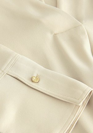 Beige pluus, millel on sile tekstuur, kuldkirsi aktsent ja peened õmbluse detailid kaeluses ja varrukas.