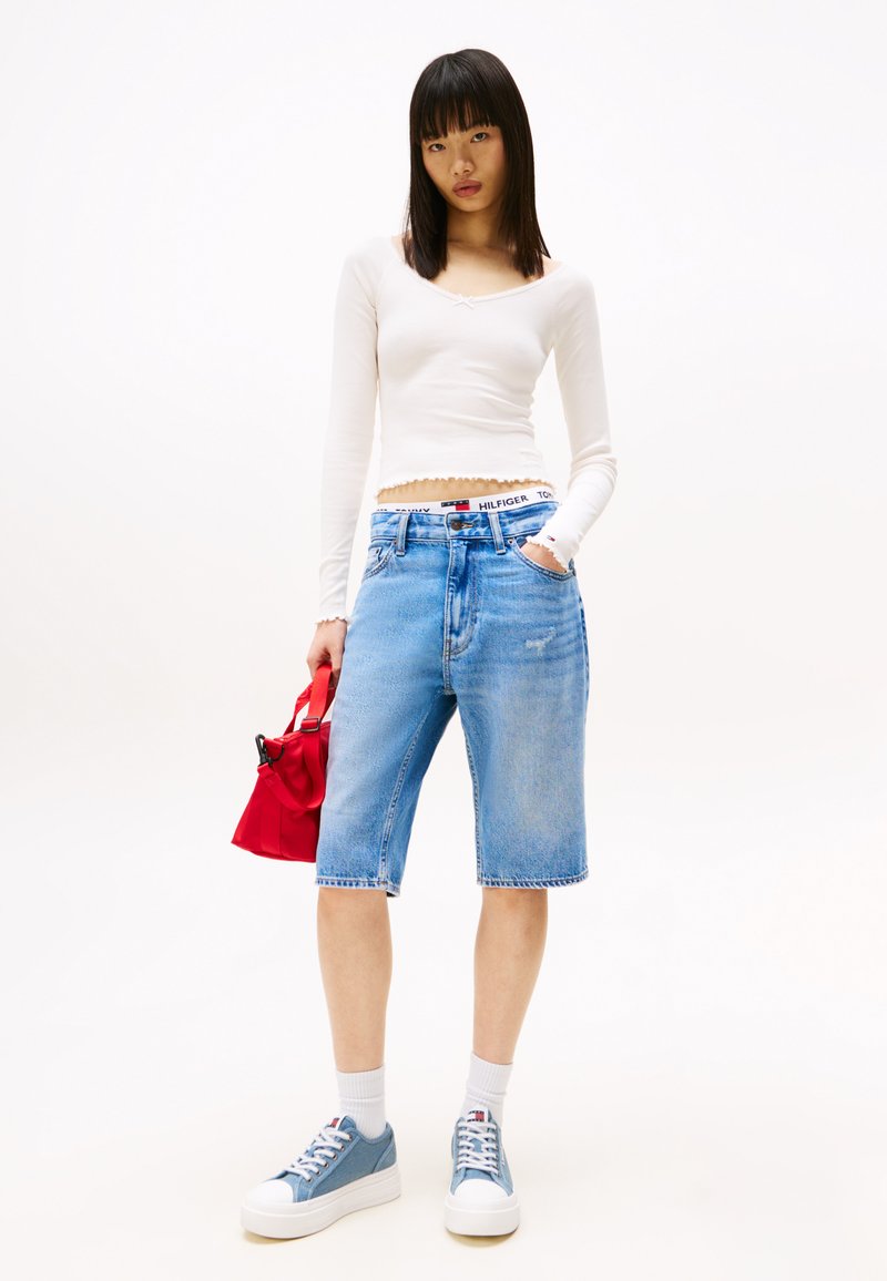 Leichtes langärmliges Oberteil, weiß; blaue Denim-Shorts mit leichter Abnutzung; rote Handtasche; blaue Sneaker mit weißen Sohlen; strukturiertem Stoff.