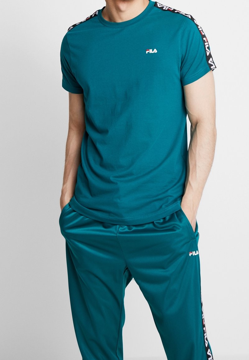 Homme portant un t-shirt FILA à manches courtes bleu sarcelle et un pantalon de survêtement assorti, les mains dans les poches, sur un fond uni.