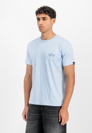 Junger Mann trägt ein hellblaues T-Shirt mit kurzen Ärmeln und kleinem Stern-Logo auf der Brust sowie dunkelgraue Jeans, steht vor weißem Hintergrund.
