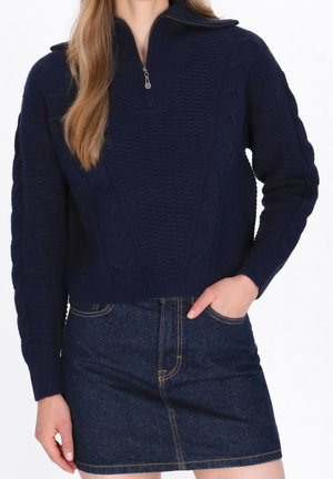 Femme portant un pull en maille torsadée bleu marine avec un col à demi-fermeture éclair et une jupe en jean foncé avec des surpiqûres contrastantes.