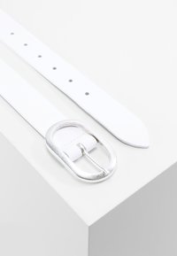 Hvid læderbælte med en sølvfarvet oval spænde. Har jævnt fordelte huller langs remmen og en tilspidset ende for et slankt design.