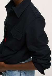 Personne portant une chemise foncée boutonnée avec les manches retroussées et un jean bleu clair, montrée du milieu du torse à l'épaule.