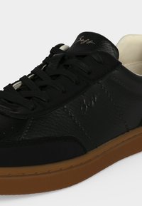 Baskets basses en cuir noir avec lacets noirs, semelle en caoutchouc marron et doublure intérieure beige, présentant un logo de marque embossé sur le côté.