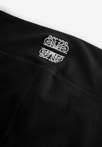 Fekete anyag sima textúrával, melyen részletes fehér hímzett minta látható. A varrás az illesztés mentén tiszta befejezést kölcsönöz.