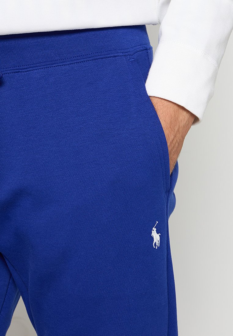 Prawa ręka w kieszeni królewskich niebieskich dresów z białym logo polo, w połączeniu z białą bluzką z długim rękawem.