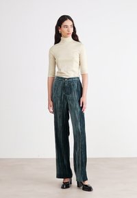 Un turtleneck doré à côtes et à manches longues associé à un pantalon large plissé vert foncé avec des rayures verticales et une fermeture à bouton. Des chaussures noires.