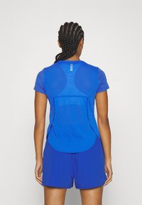 Blå idrottstopp med korta ärmar, texturerad tyg och andningsbart nättyg. Har en rundad nederkant och subtila reflektionsdetaljer.