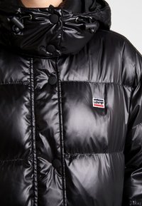 Černá prošívaná bunda s lesklým, prošitým povrchem, vybavená vysokým límcem, patentními knoflíky a malým vyšívaným logem Levi's na hrudi.