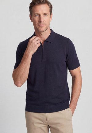 Mann trägt ein marineblaues kurzärmeliges Polohemd mit Halbreißverschluss-Kragen und beige Hose, steht vor einem einfarbigen hellen Hintergrund.