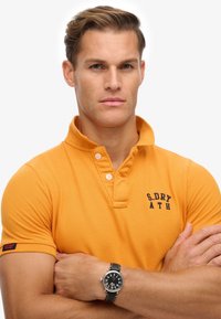 Oranje polo shirt met een kraag en twee knopen, met een marineblauw logo. Het model draagt een zwart horloge met een leren band.