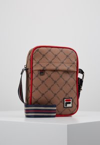 Sac synthétique marron avec un motif de logo répétitif, garniture rouge et une bandoulière ajustable rayée. Il dispose d'une poche avant avec un détail de logo.