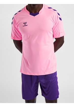 Athlète portant un maillot de sport rose avec des bandes chevron violettes sur les épaules et un short violet, debout les mains dans le dos.