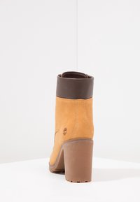 Bottine en daim camel avec un revers en cuir brun foncé, un talon haut et une semelle en caoutchouc texturée. Présente un logo embossé sur le côté.