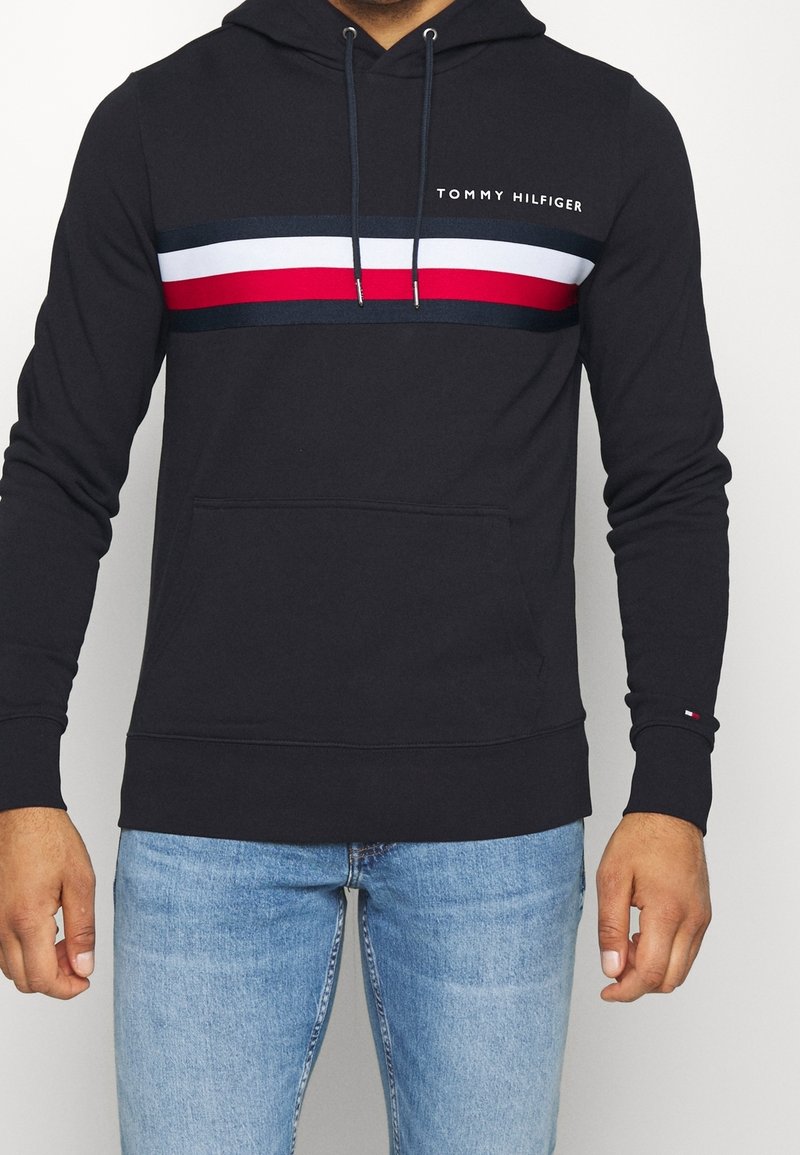 Hoodie bleu marine avec une poche frontale, présentant des rayures horizontales rouge, blanc et bleu, et le logo "TOMMY HILFIGER" brodé sur la poitrine.