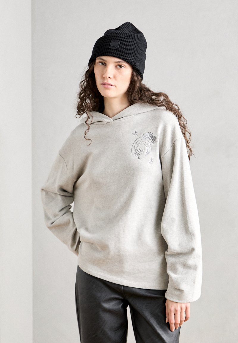 Felpa grigia chiara con tasca frontale e design grafico, abbinata a un berretto a coste nero. Modello che indossa pantaloni in similpelle nera.