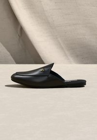 Μαύρο δερμάτινο mule με κατευθείαν μύτη και μικρή χρυσή διακόσμηση σε σχήμα καρδιάς, σε ελαφρώς μπεζ επιφάνεια με υφασμάτινο παρασκήνιο.