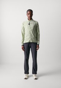 Veste zippée à manches longues vert clair, avec une texture lisse, dotée de poches latérales et d'accents noirs ; associée à un pantalon gris foncé et des chaussures légères.