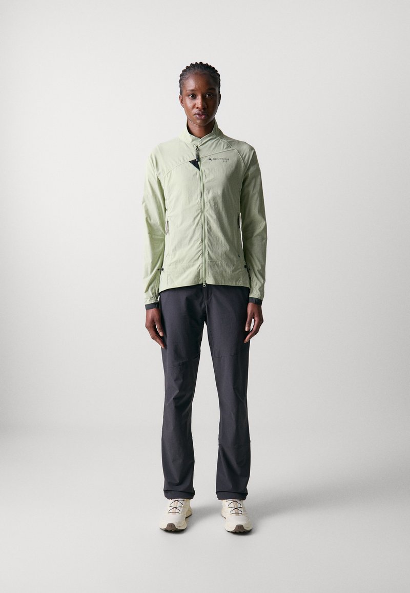 Veste zippée à manches longues vert clair, avec une texture lisse, dotée de poches latérales et d'accents noirs ; associée à un pantalon gris foncé et des chaussures légères.
