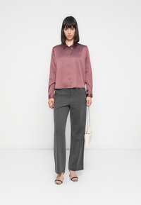Blusa in satin rosa con colletto e chiusura a bottoni, abbinata a pantaloni grigi tailored. Gli accessori includono una piccola borsa bianca e sandali multicolori.