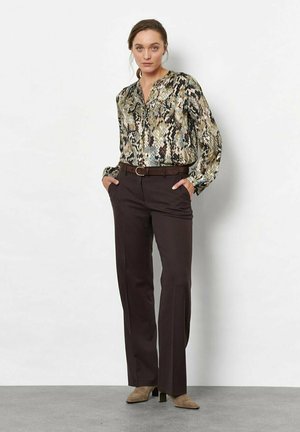 Donna in piedi con le mani nelle tasche, indossa una camicetta a maniche lunghe con stampa a serpente, pantaloni marrone scuro, cintura marrone e scarpe beige con tacco.