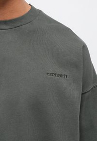 Tume roheline dressipluus ribidega kaeluses, millel on rinnal peenelt broderitud "carhartt" logo, valmistatud tekstuurilisest kangast.