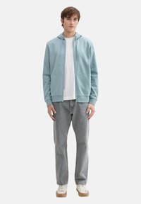Svetlomodrá mikina na zips s predným vreckom, kombinovaná s bielym tričkom a svetlosivými džínami. Model má na sebe biele tenisky s béžovými podrážkami.