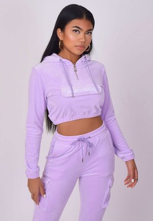 Donna con capelli lunghi e scuri, indossa orecchini a cerchio dorati, una felpa corta lavanda e pantaloni lavanda coordinati con coulisse e tasche cargo.