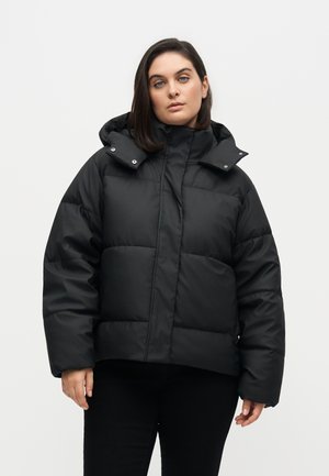 Mujer con cabello liso y oscuro que lleva una chaqueta acolchada negra con cuello alto y capucha, de pie frente a un fondo claro y liso.