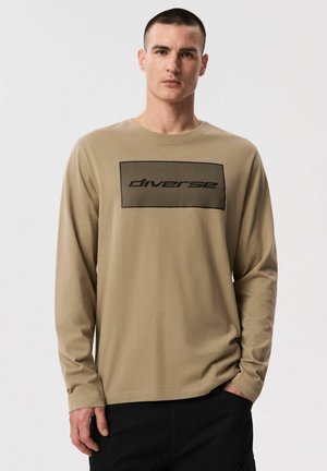 Mężczyzna ubrany w beżową koszulę z długim rękawem z czarnym prostokątnym nadrukiem i tekstem, w czarnych spodniach, stojący na białym tle.