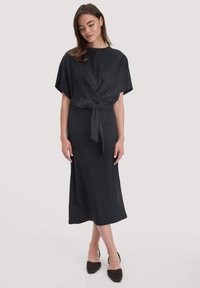 Frau in einem schwarzen Midikleid mit kurzen Ärmeln und einer gebundenen Taillen-Schleife, kombiniert mit schwarzen spitzen Slingback-Schuhen.