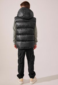 Chaleco de plumas negro con capucha, que presenta una textura brillante y un patrón acolchado. Lleva puesto sobre una camiseta gris de manga larga y se combina con pantalones cargo negros.