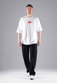 Weißes übergroßes T-Shirt mit einem roten Logo-Patch, kombiniert mit lockeren schwarzen Hosen und weißen Sneakers. Schlichte Gestaltung, aus Baumwollmaterial.